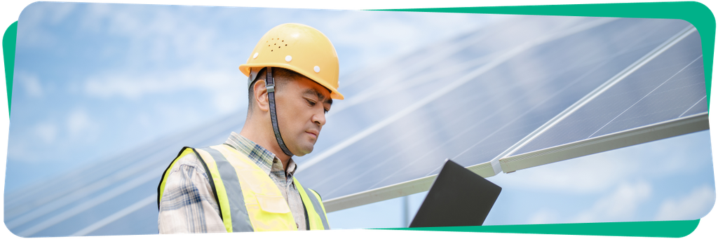 A man in a hard hat and safety vest inspects solar panels while holding a tablet, with a blue sky background.