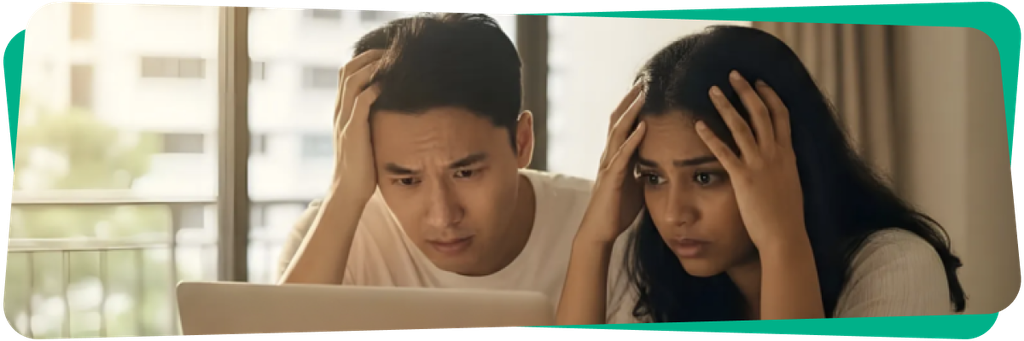 A worried couple looks at a laptop screen, sitting indoors with hands on their heads, by a window with soft lighting.