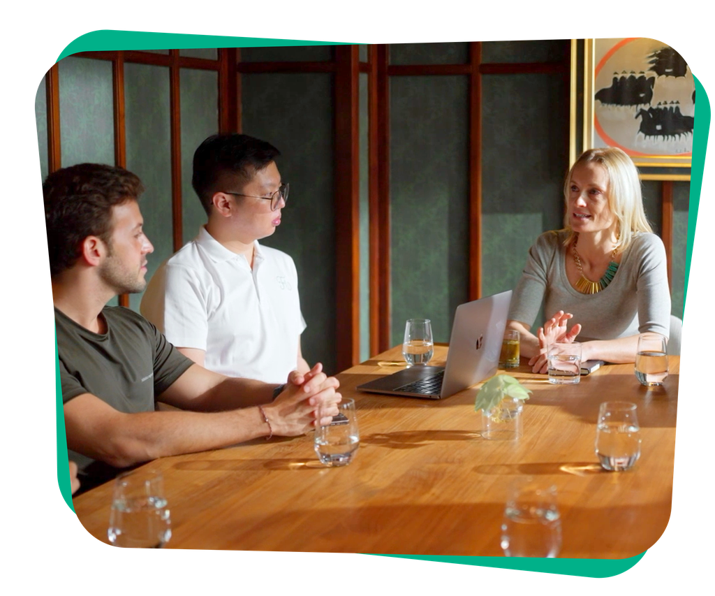 Three colleagues seated around a wooden conference table, a woman speaks beside a laptop while two men listen; glasses of water on the table.