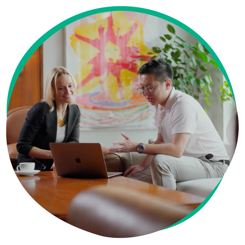 Two colleagues seated on a sofa reviewing a laptop on a wooden table, coffee cup nearby, colorful painting and plant in the background.